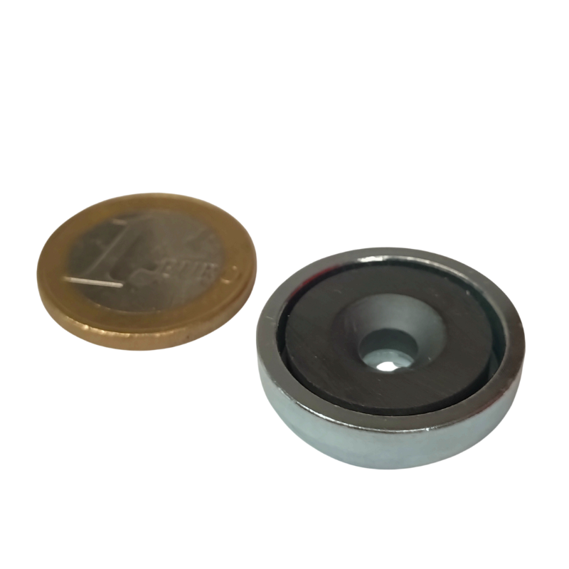 Een muntstuk van een euro ligt naast een Keramische potmagneet met verzonken gat 25mm hoogte 7mm Houdkracht 3kg, beide geplaatst op een witte achtergrond.